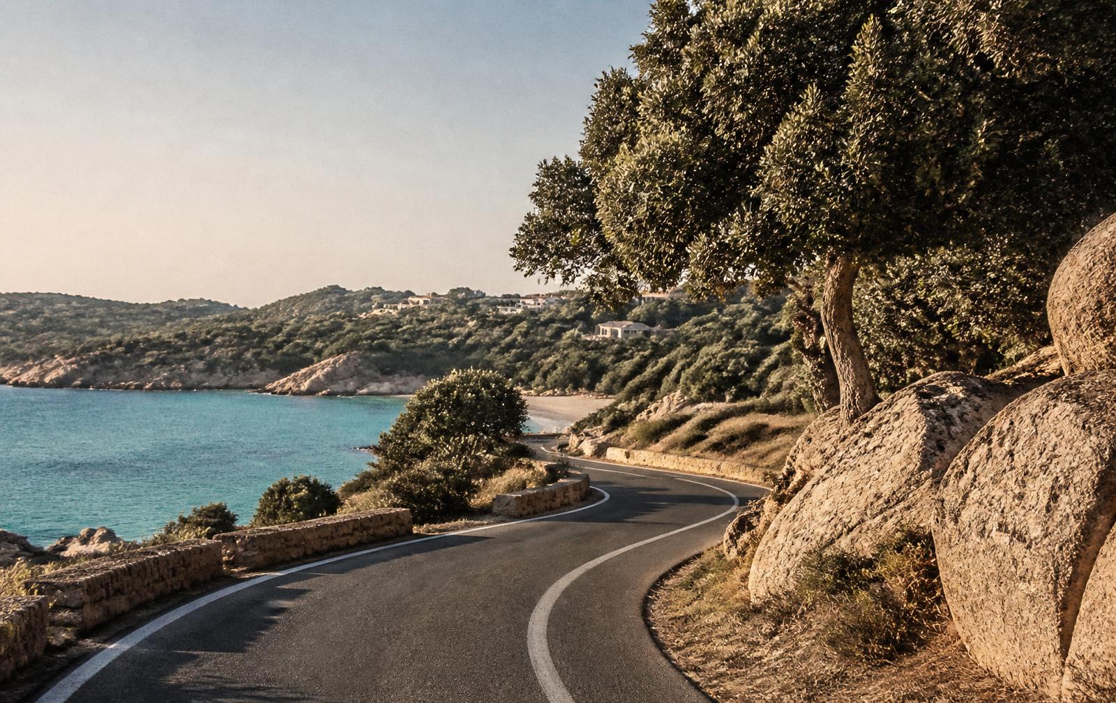 Early morning on an empty Gallura coast road curving between granite boulders toward a turquoise bay
